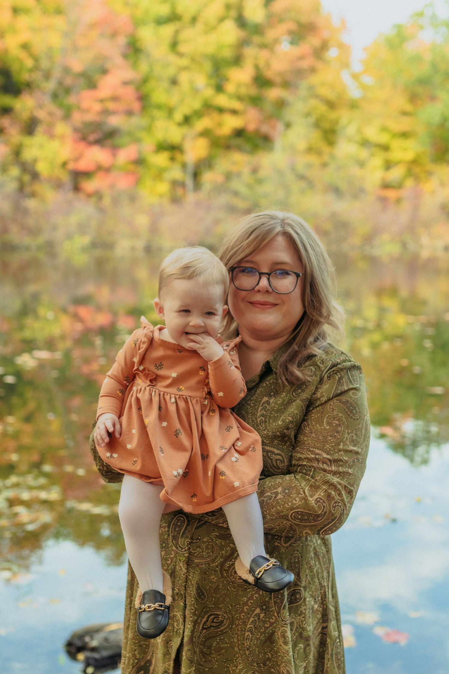 A parent and child share a tender moment outdoors against a backdrop of autumn foliage and reflective water.