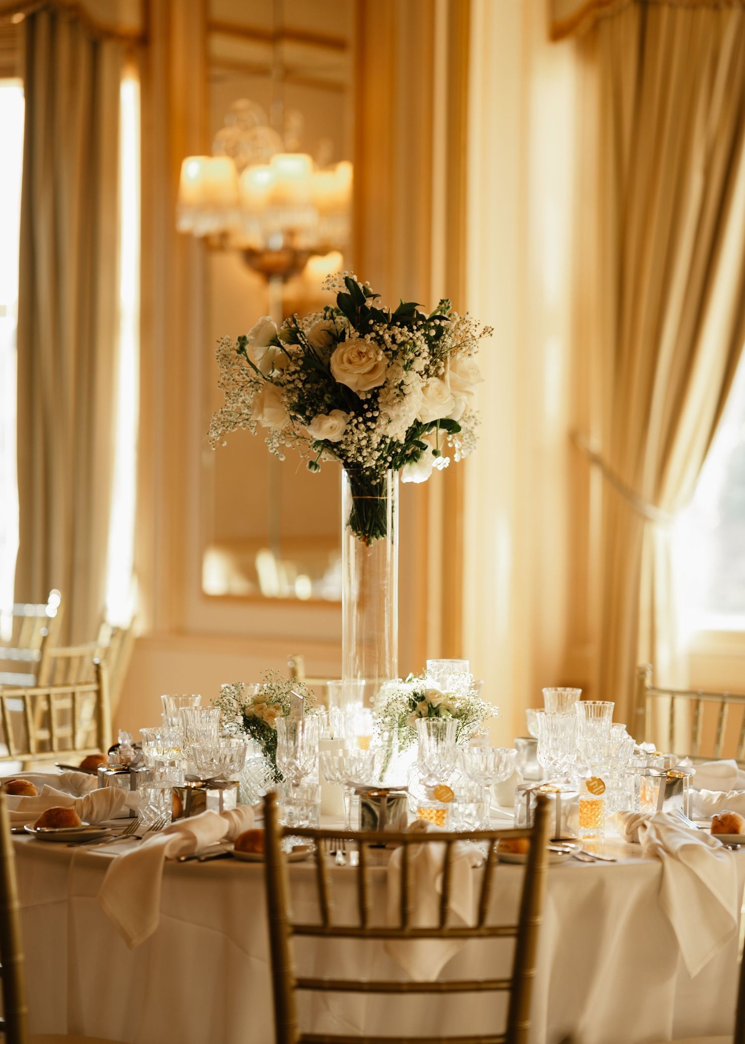 Elegant wedding reception table with tall white floral centerpiece and formal place settings in a golden-lit ballroom.