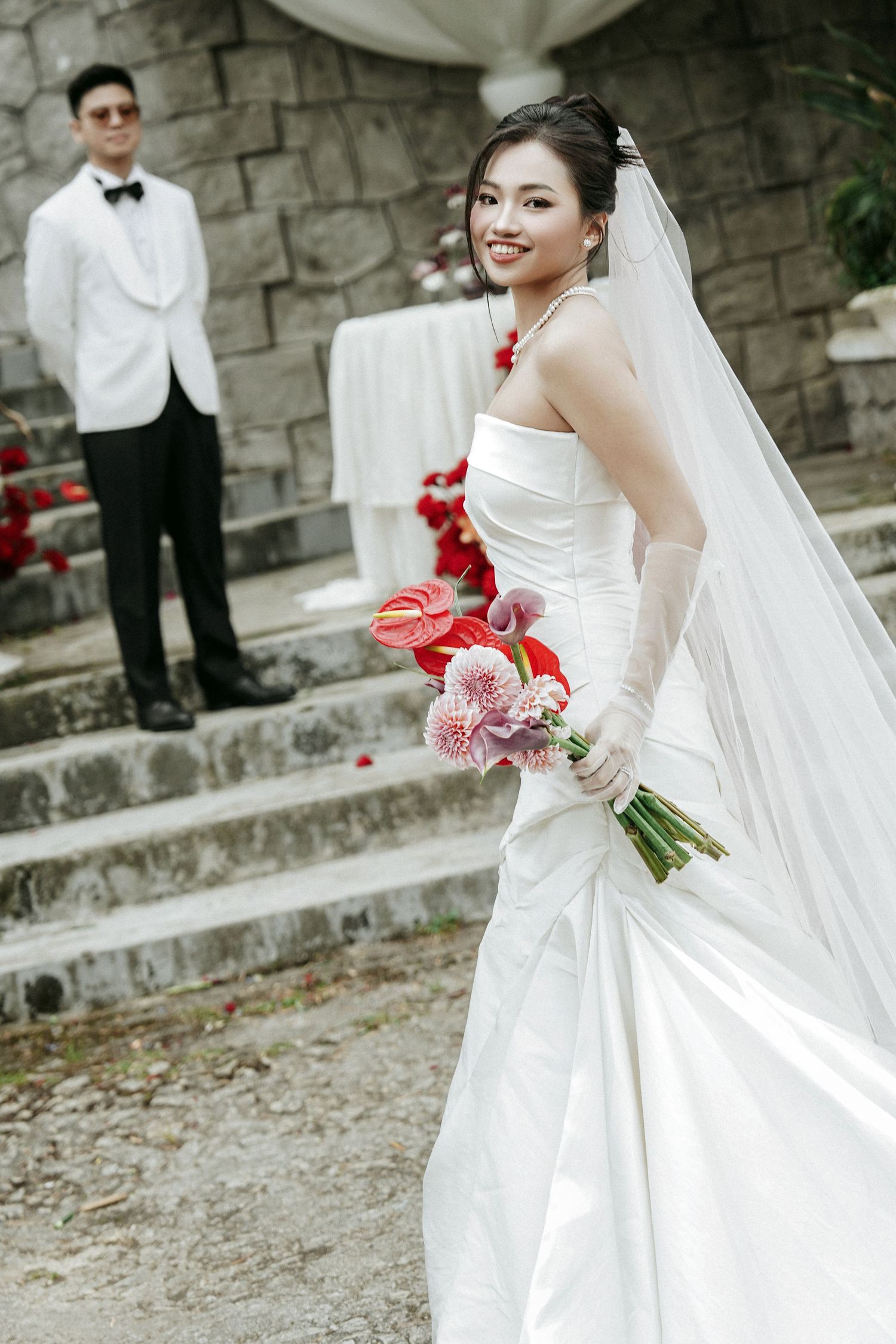 Smiling Asian bride in strapless white gown holding floral bouquet with groom standing on stone steps behind her.