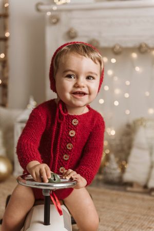 Toddler in red knit cardigan and hood sitting on stool with festive holiday backdrop.