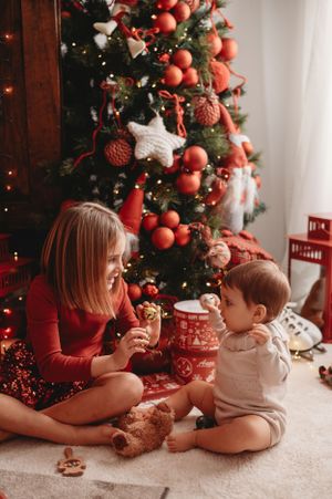 Two children sharing Christmas treats in front of a decorated tree with red ornaments.