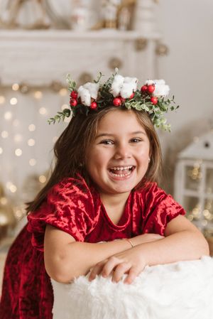 A festive portrait with a red velvet dress and white floral crown with berries against twinkling lights.