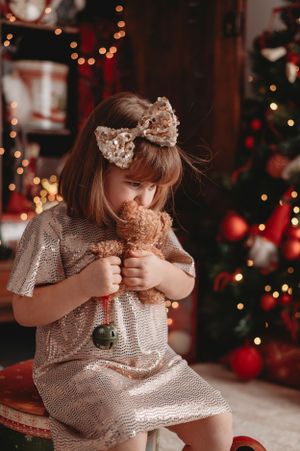 Small child in sparkly dress holding teddy bear in front of Christmas tree with red ornaments.