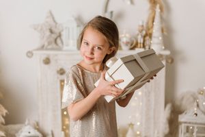Child in sparkly dress holds white gift box in front of white holiday mantel.