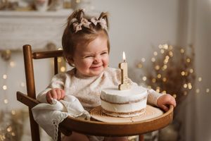 Toddler sits at highchair with small birthday cake and single candle against holiday lights.