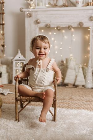 Toddler in cream romper sits happily on wooden chair against white holiday backdrop.