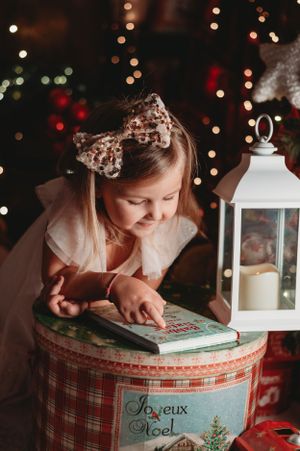 Child reading a book on a Christmas-themed box near a white lantern with twinkling holiday lights in the background.