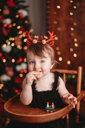 Child wearing reindeer antlers sitting at table with toy train and Christmas lights in background.