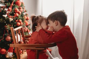 Two children in red sweaters sharing a secret in front of decorated Christmas tree.