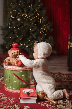 A baby in white knit outfit reaches for teddy bear on Christmas decorated container near tree.