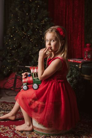 A thoughtful holiday portrait in red dress beside vintage toy train and decorated Christmas tree.