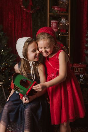Two children share a playful moment in knit hats during a festive Christmas photoshoot with red decor.