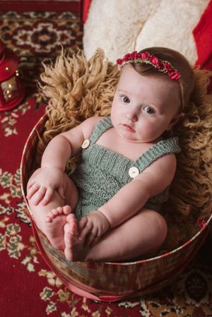 A baby wearing mint green knit romper sits in wooden basket on red and beige patterned rug.