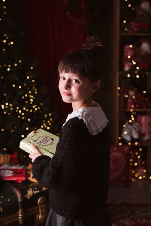 Child in vintage-style outfit holds book against Christmas backdrop with twinkle lights.
