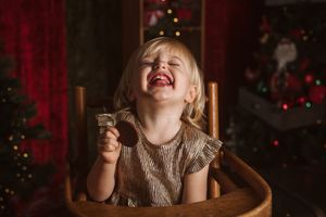 A candid holiday portrait showing pure joy while sitting in wooden chair with Christmas decor.