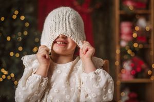 Child playfully pulls knit winter hat over face against Christmas lights background.