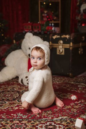 Small child in white knit outfit sits on patterned rug with Christmas decorations and teddy bear.