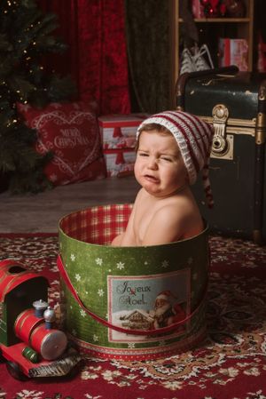 Baby wearing striped winter hat sits in festive green Christmas tin container.
