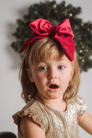 Young child wearing gold dress and large red bow headband poses against holiday background.