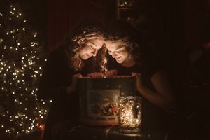 Two figures peer into a glowing lantern in a dark room with Christmas lights in the background.