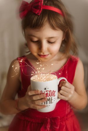 Young child in red dress and bow holding a festive winter mug with sparkles.