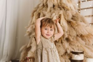 Festive holiday scene with child in beige dress raising arms by decorated tree.