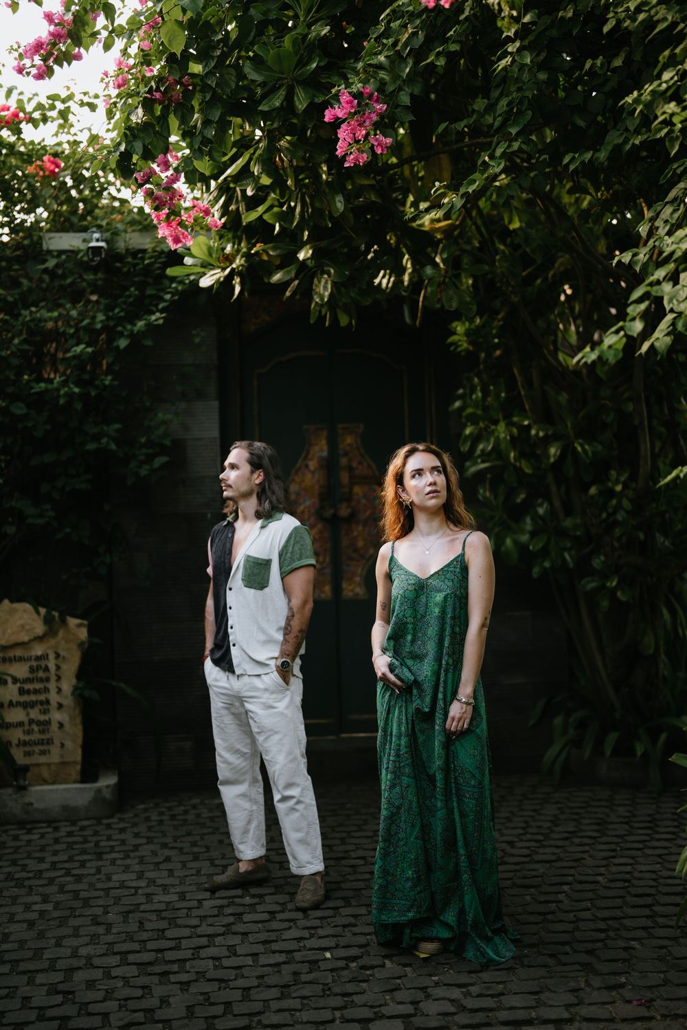 A couple poses against a dark garden wall with pink bougainvillea flowers overhead.