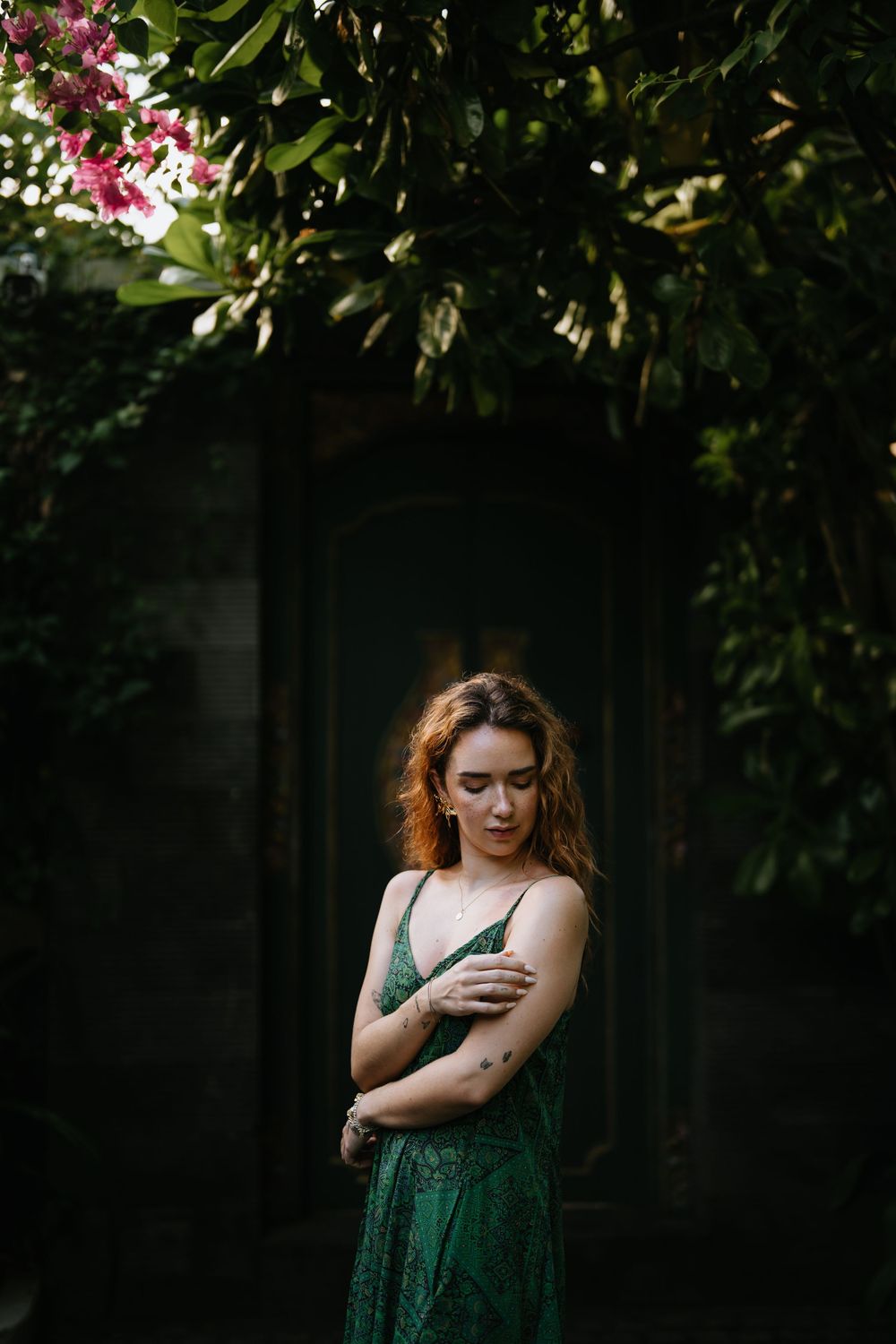 A person in a green dress stands thoughtfully in a garden with pink flowers and dark foliage.