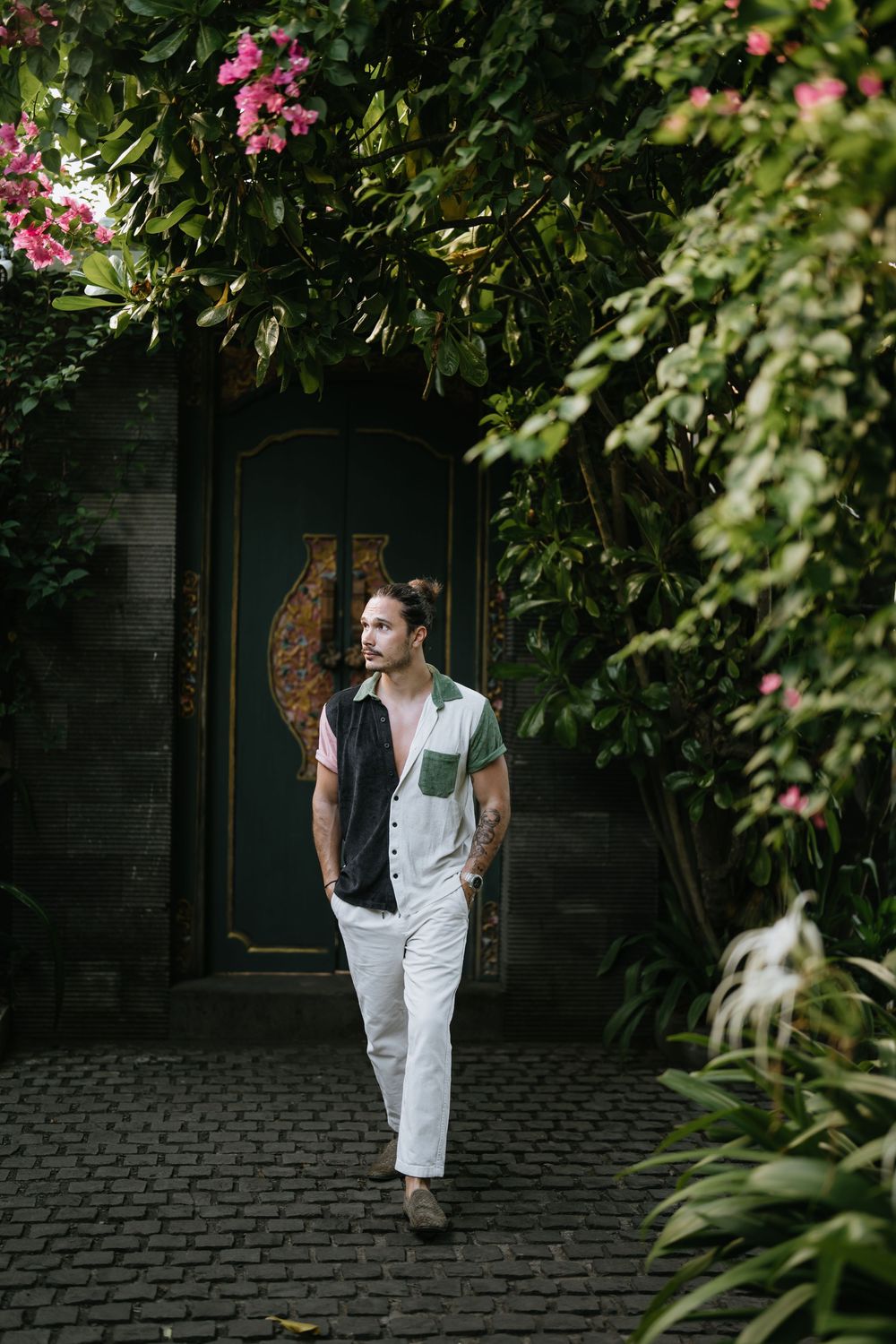 Figure in white clothing poses against lush green foliage and pink flowers in a garden setting.