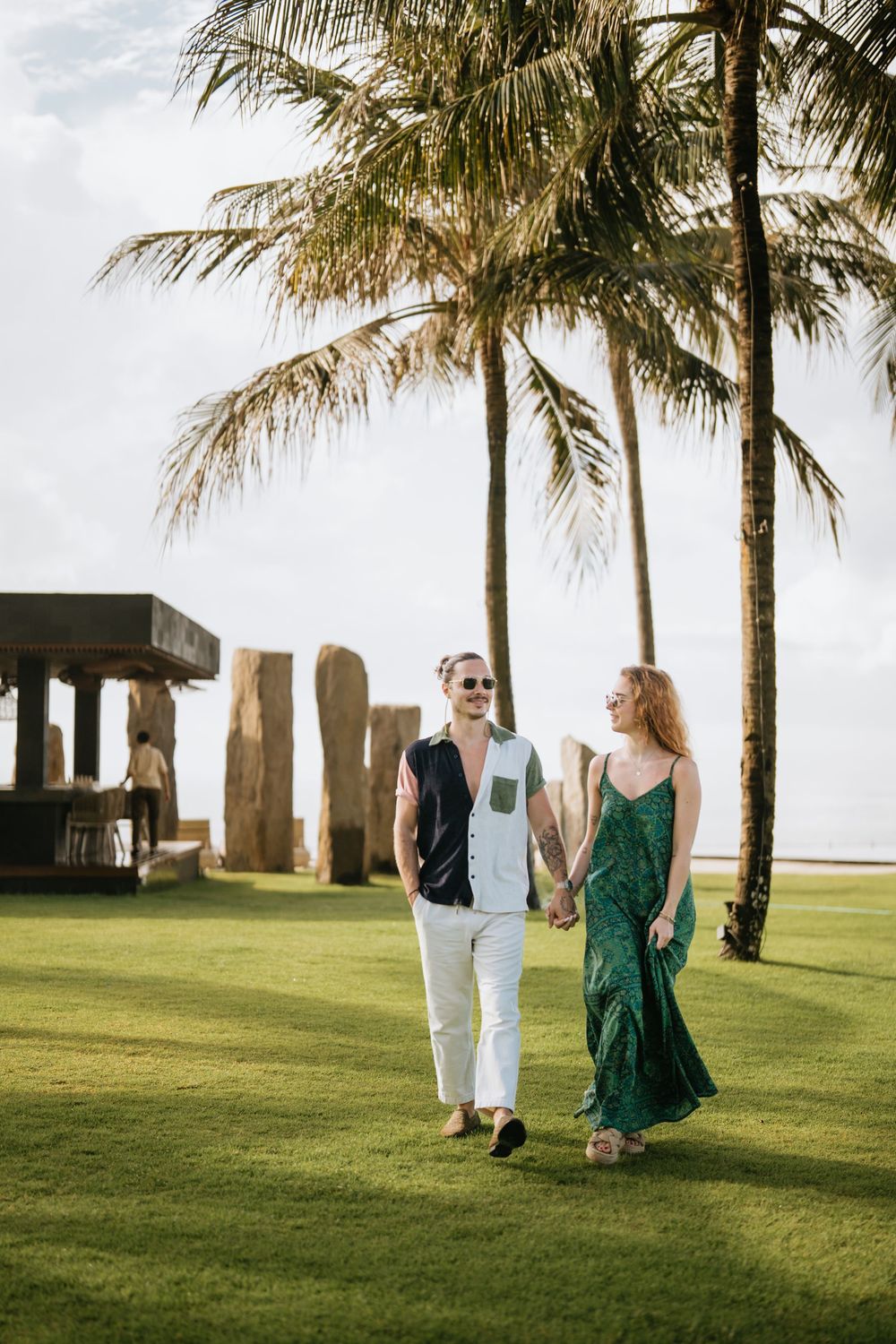 Couple walking along a grassy beachfront path with palm trees swaying in the breeze.