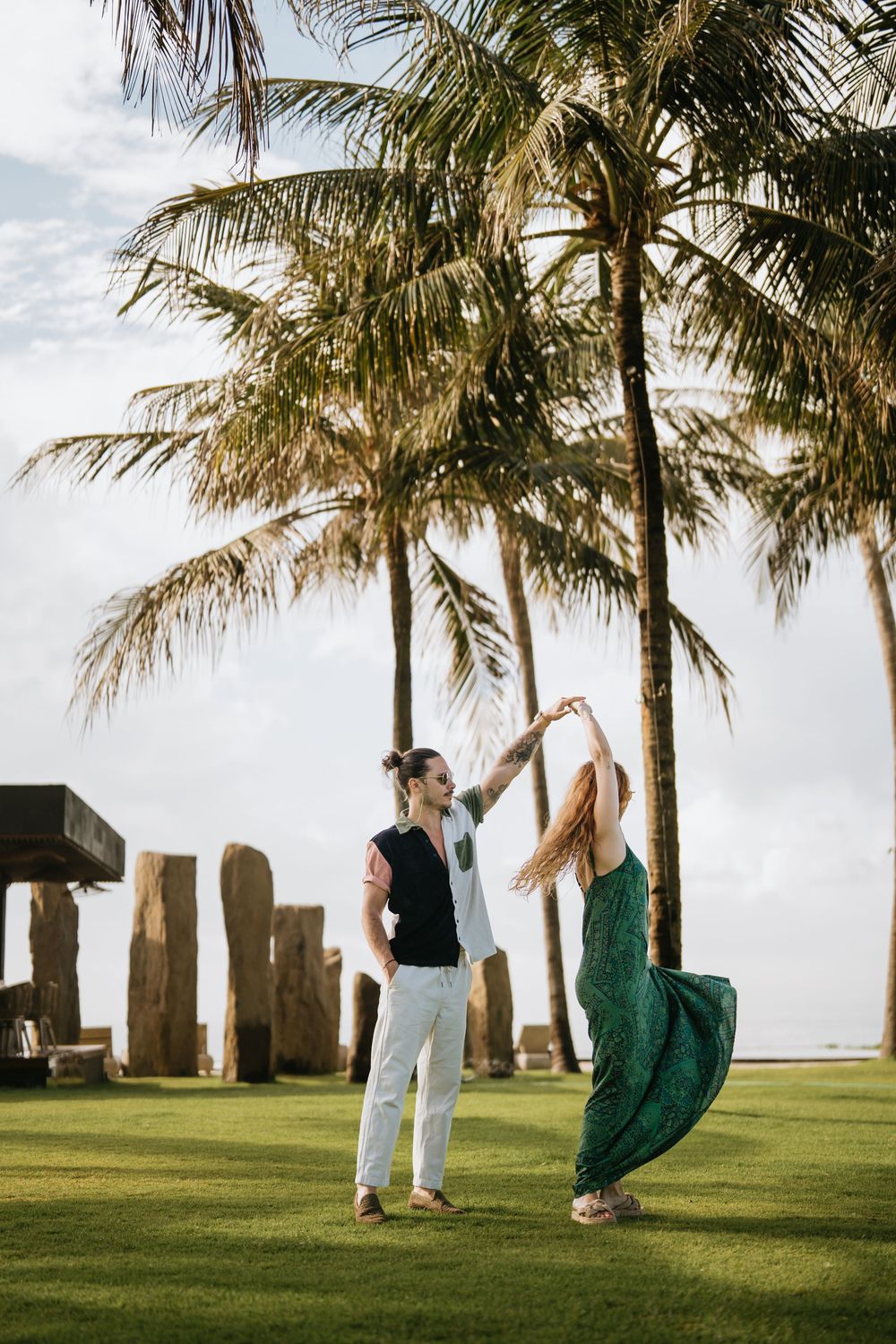 Two people dance playfully on green grass with palm trees and stone structures in the background.