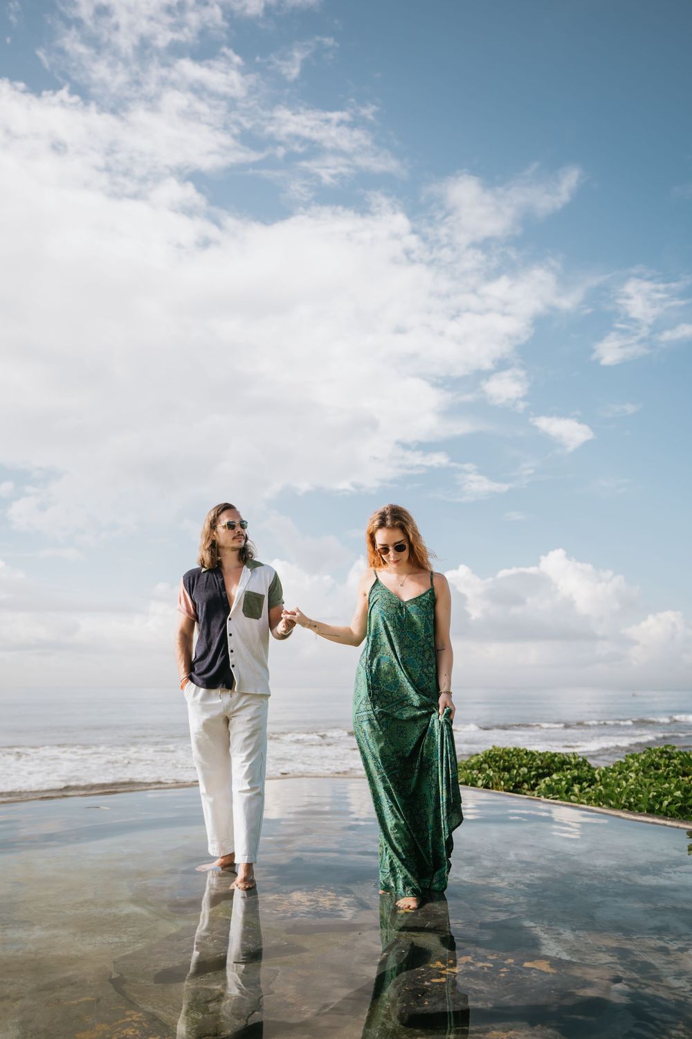 Two people stand together on a beach with ocean waves and cloudy sky in the background.