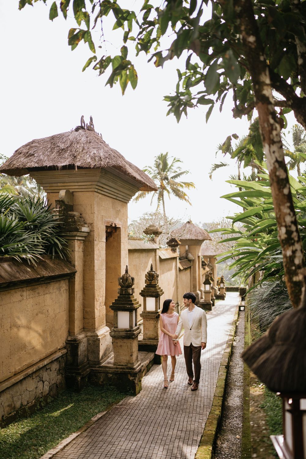 A couple walks along a tropical path with traditional Balinese architecture and palm trees.