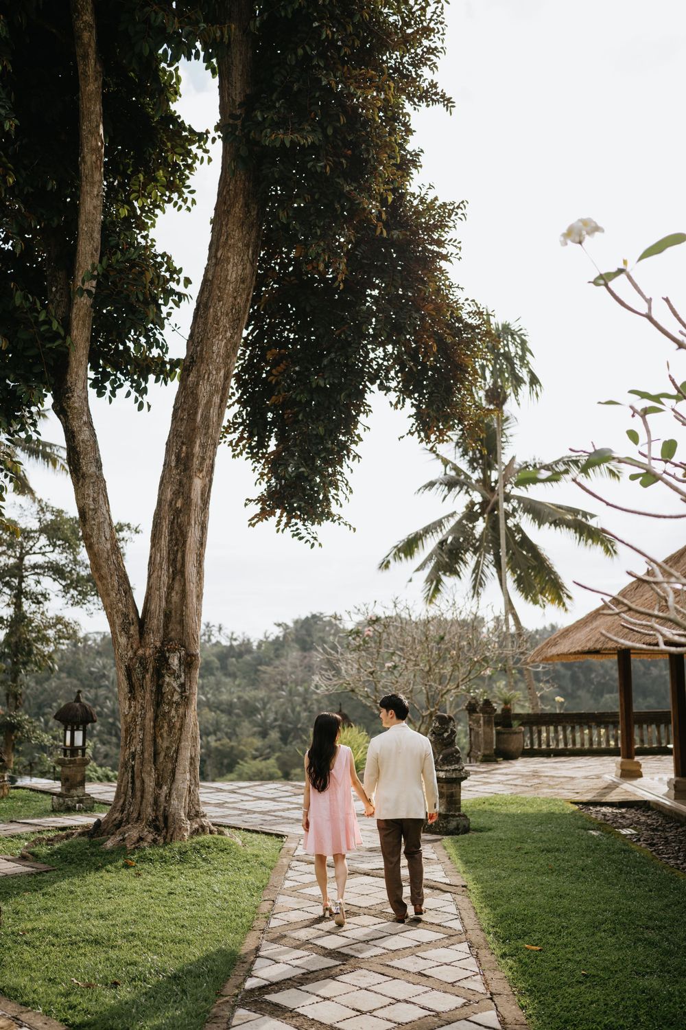 A couple walks along a scenic tropical garden path with palm trees and traditional huts.