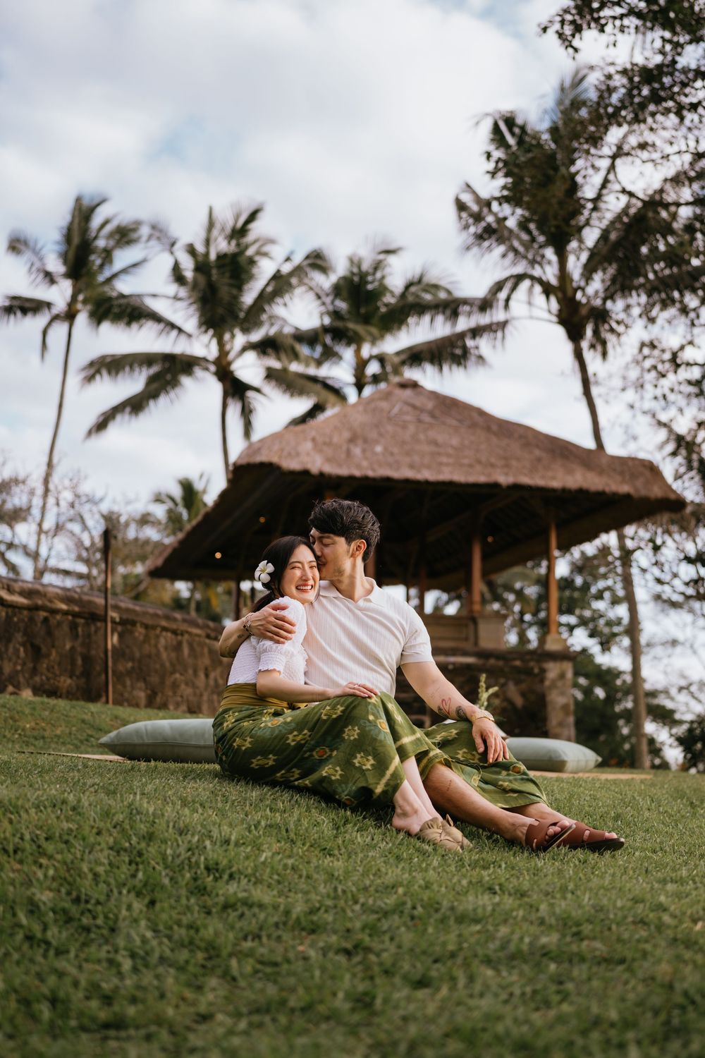 A couple relaxes on a grassy hillside with palm trees and traditional huts in the background.