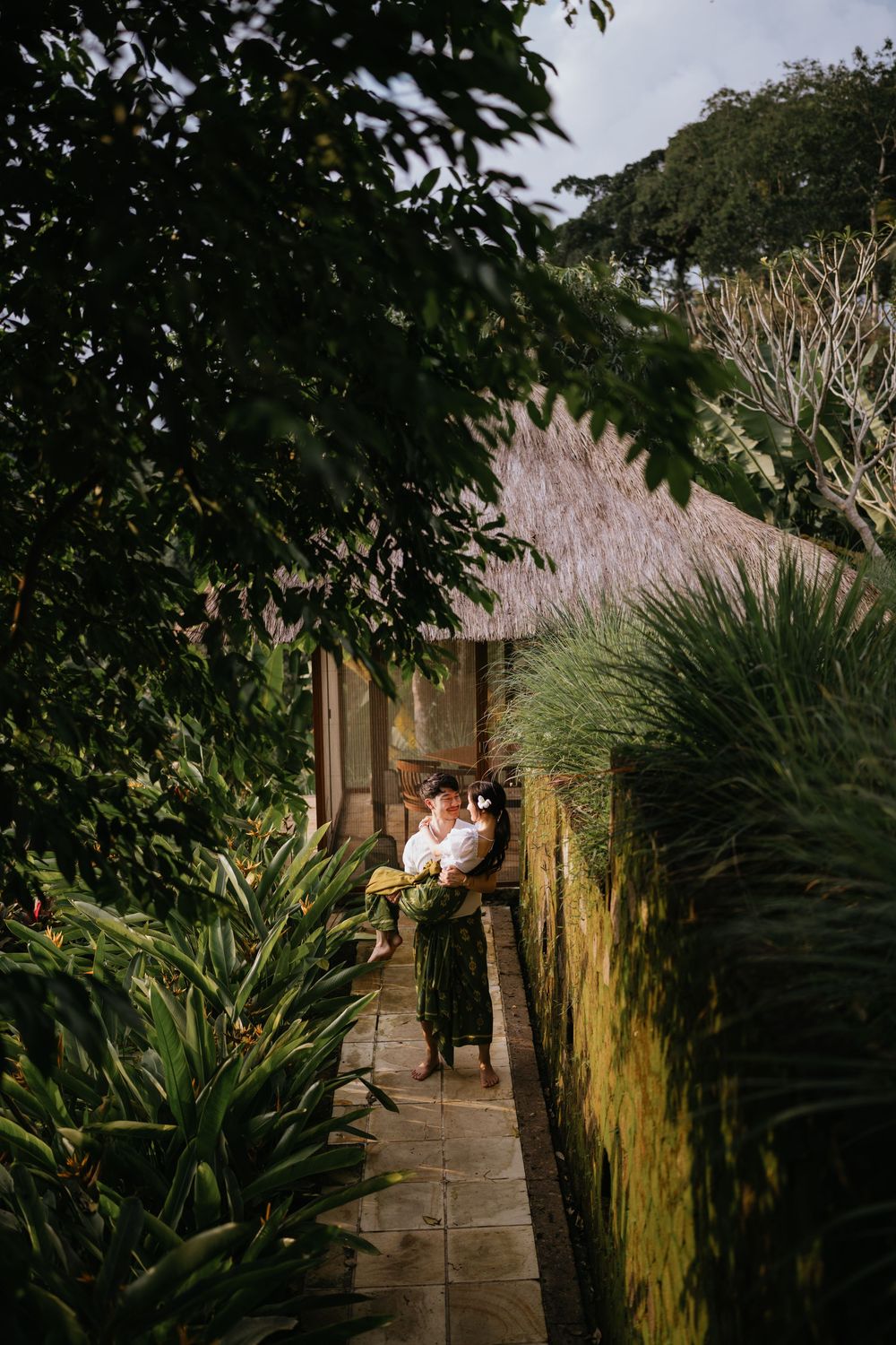 Two people share an intimate moment in a lush garden pathway with traditional architecture.