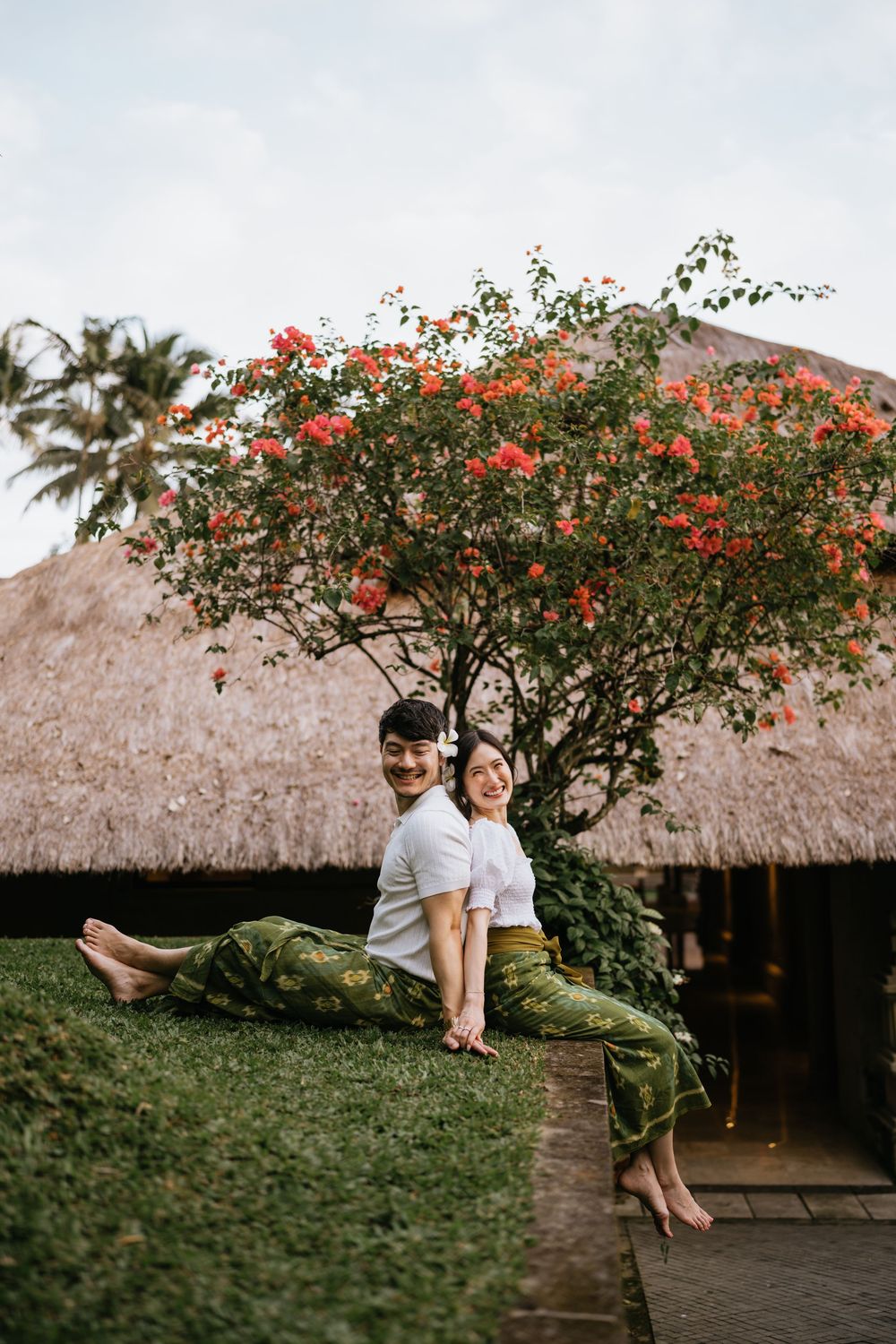 A couple relaxes on grass near a thatched roof building with vibrant red flowers in bloom.