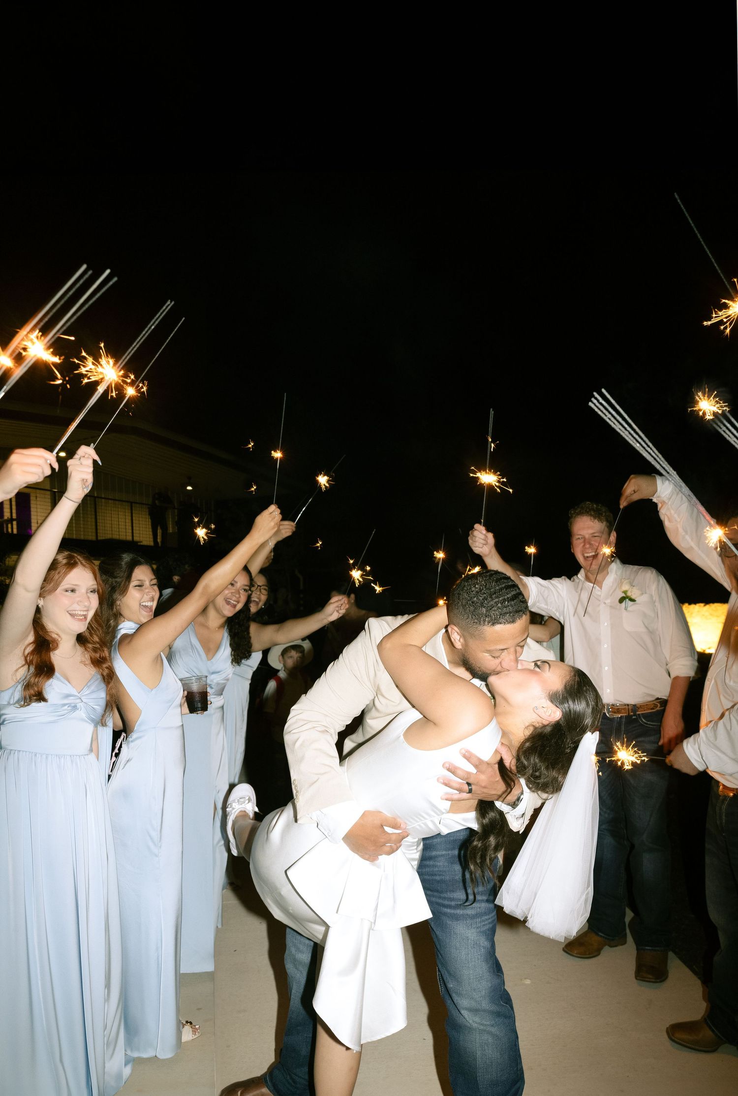 A couple shares a loving embrace during their wedding send-off while guests wave sparklers around them at night.