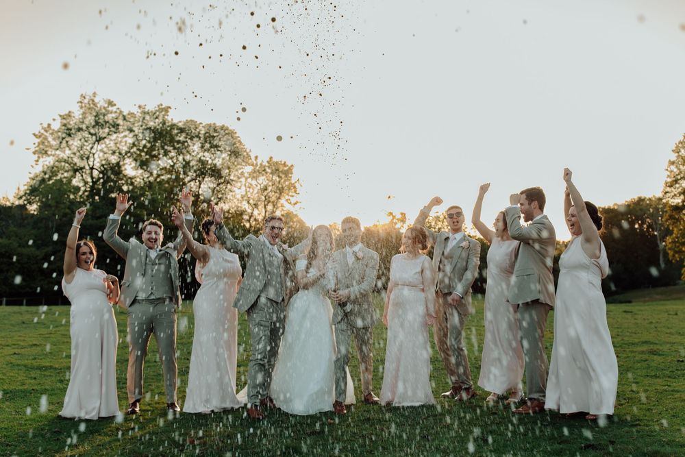 Bridesmaids in light sage green dresses celebrate and cheer on a grassy lawn during golden hour.