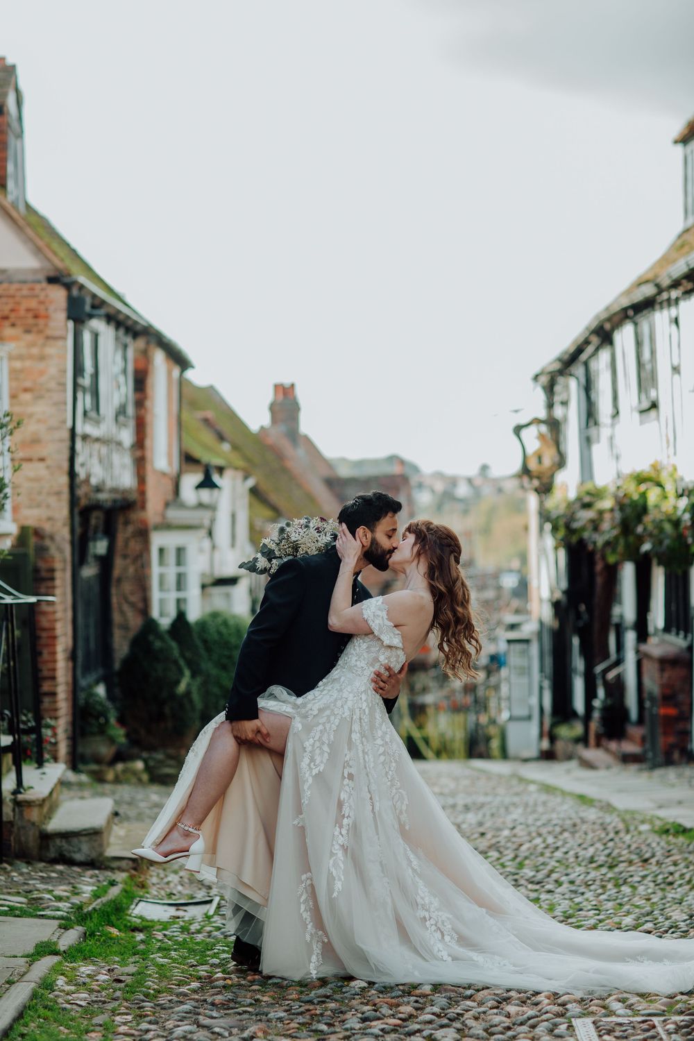 A romantic couple shares a kiss on a charming cobblestone street lined with historic buildings and greenery.