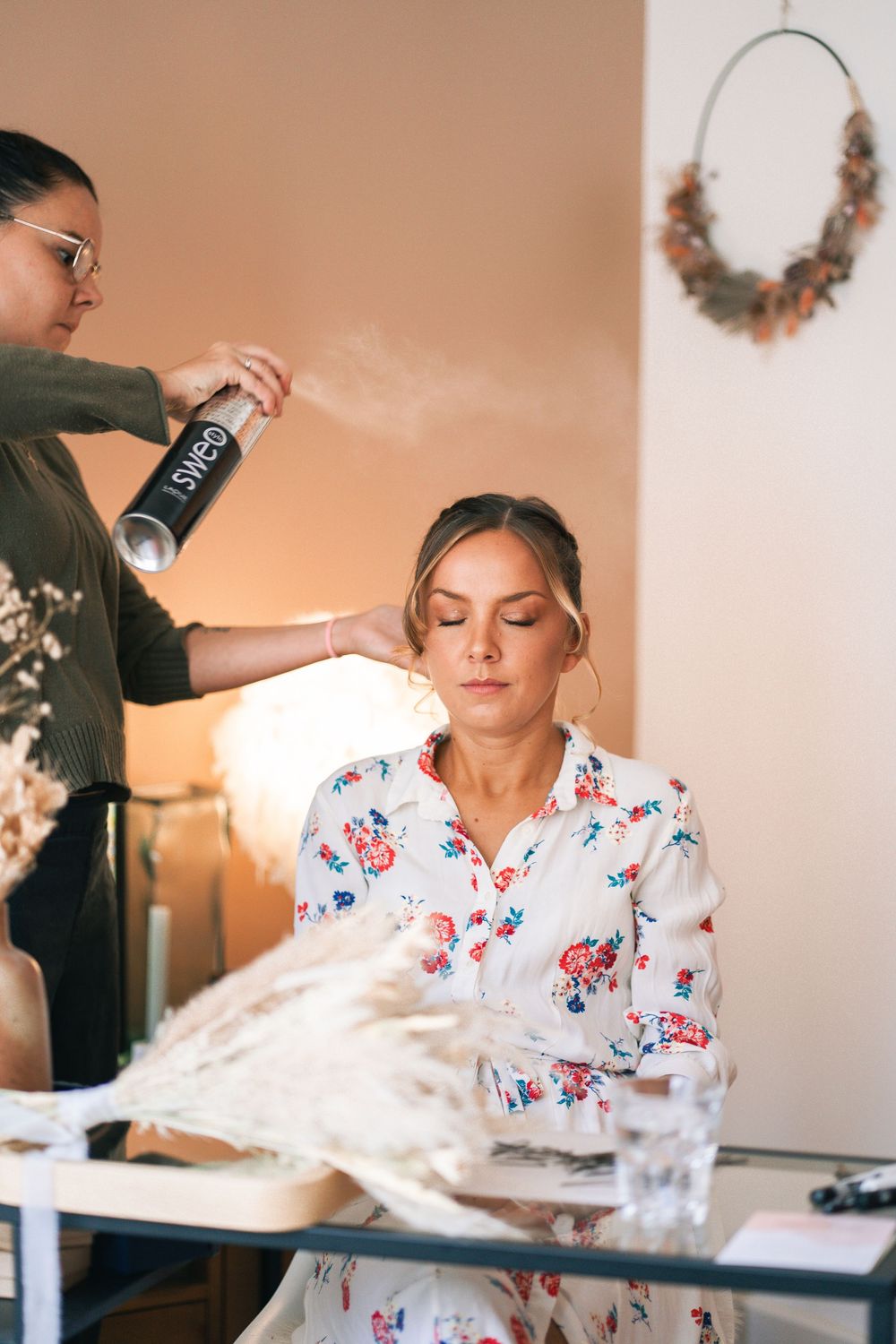 A bride getting her hair styled and makeup done while wearing a floral robe during wedding preparations.