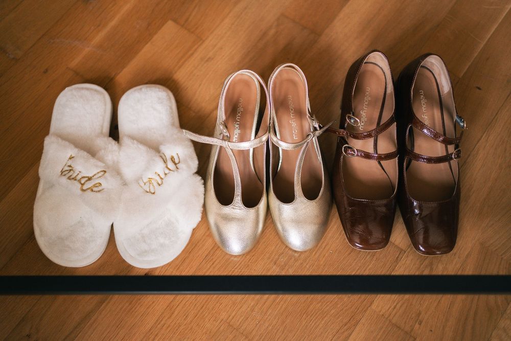 Three pairs of wedding shoes arranged on a wooden floor including white slippers, bridal heels, and brown dress shoes.