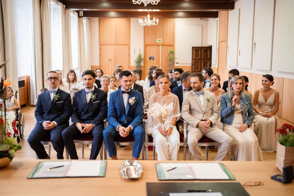 Wedding party and guests seated watching ceremony in an elegant wood-paneled room with chandelier.