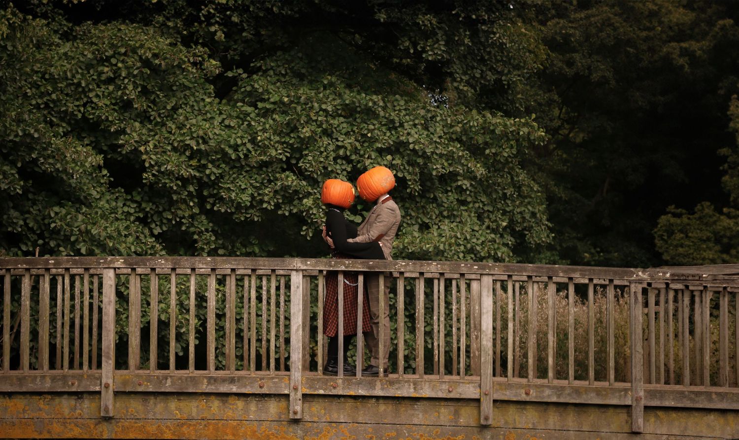 Figure with pumpkin heads embraces on a wooden bridge surrounded by autumn foliage.