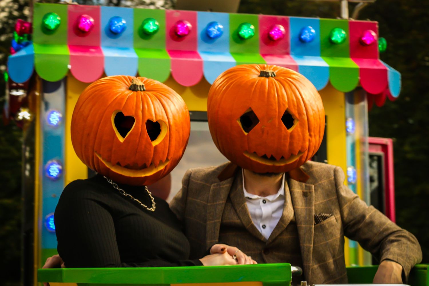 Two people wearing pumpkin heads and formal attire pose under colorful festival bunting.