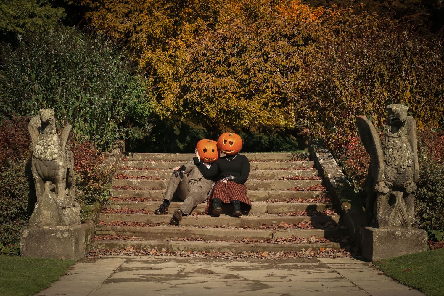 Couple with pumpkin heads posing on stone steps with lion statues on autumn day.