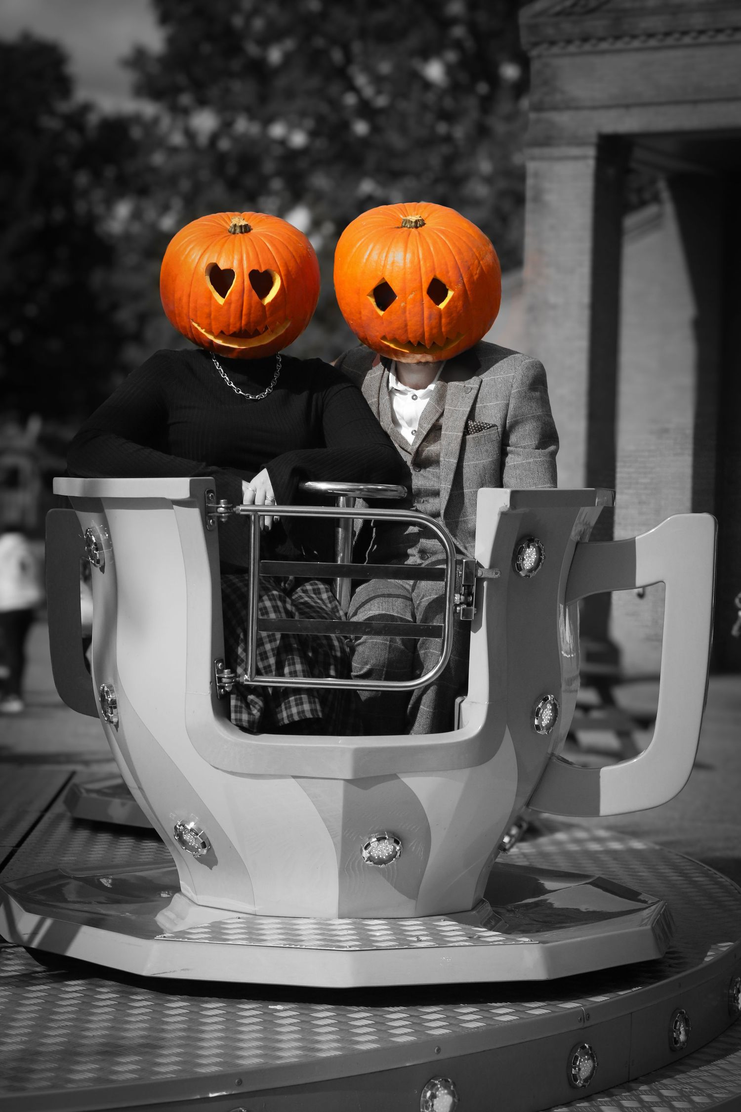 Two figures wearing pumpkin head masks ride a spinning teacup ride in a black and white photo with orange pumpkins highlighted.