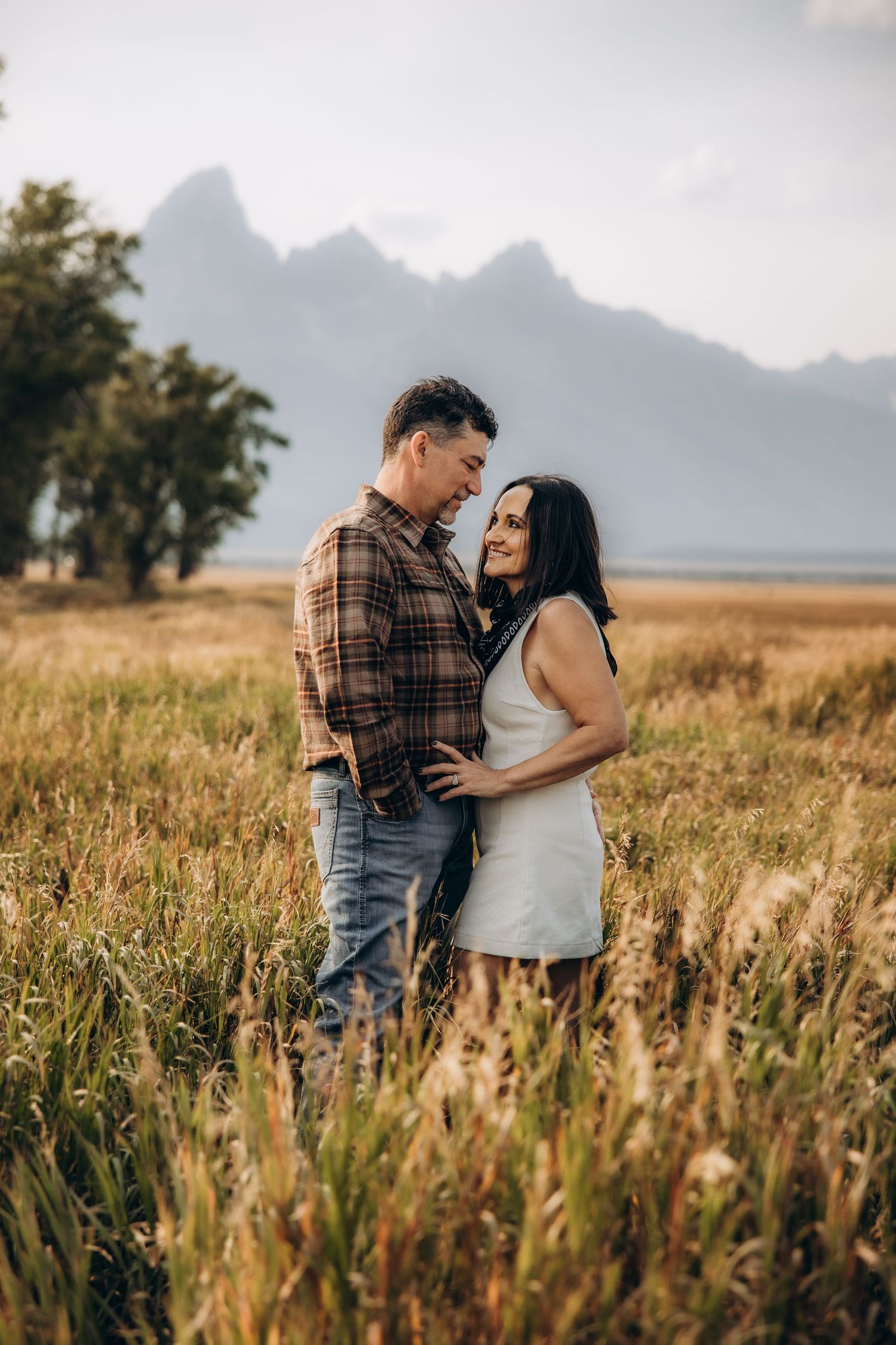A couple embraces in a golden meadow with majestic mountain peaks silhouetted against the sky in the background.
