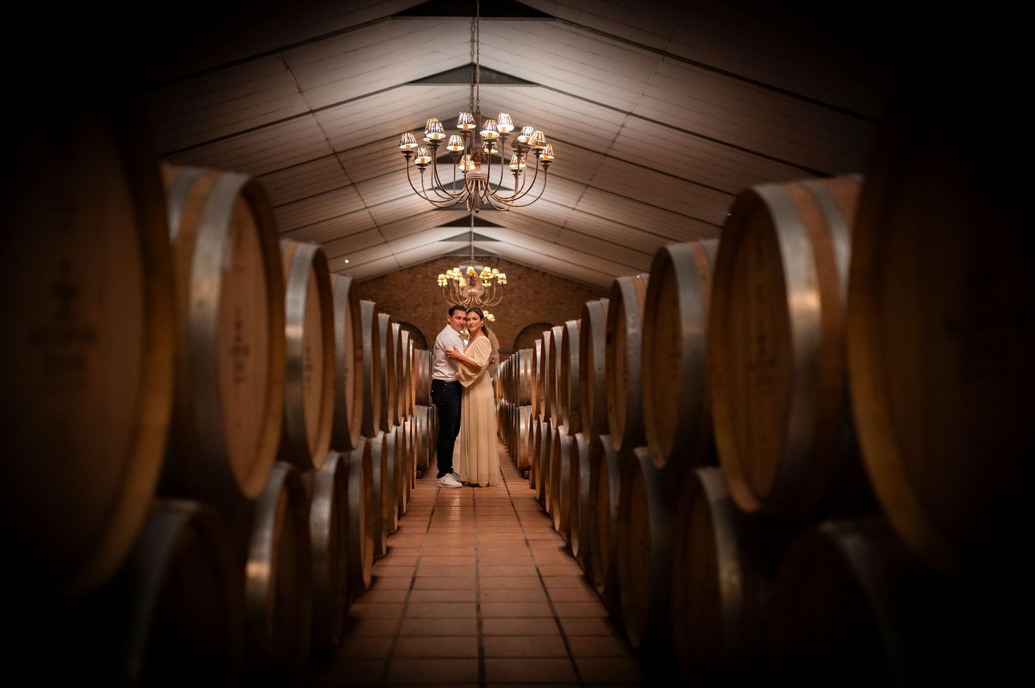 Wine barrels line the walls of an atmospheric underground cellar tunnel lit by warm chandelier lighting.