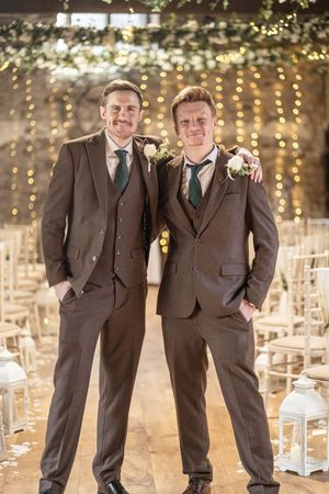 Groomsmen in brown suits pose together at an evening wedding venue decorated with twinkling lights.
